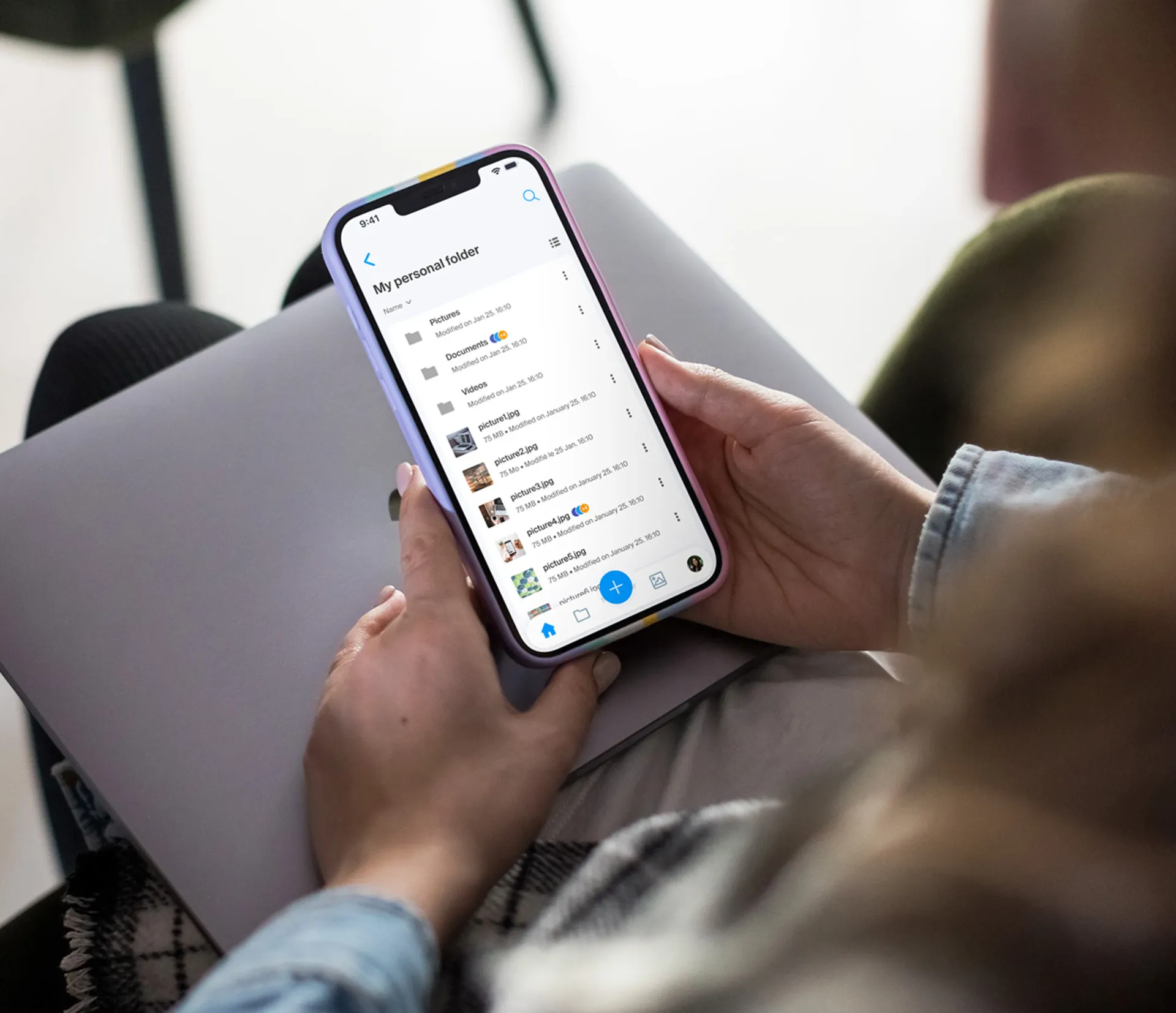 Une personne tenant un smartphone affichant une interface de gestion de fichiers sur une application mobile, avec un ordinateur portable posé sur ses genoux dans un environnement confortable.