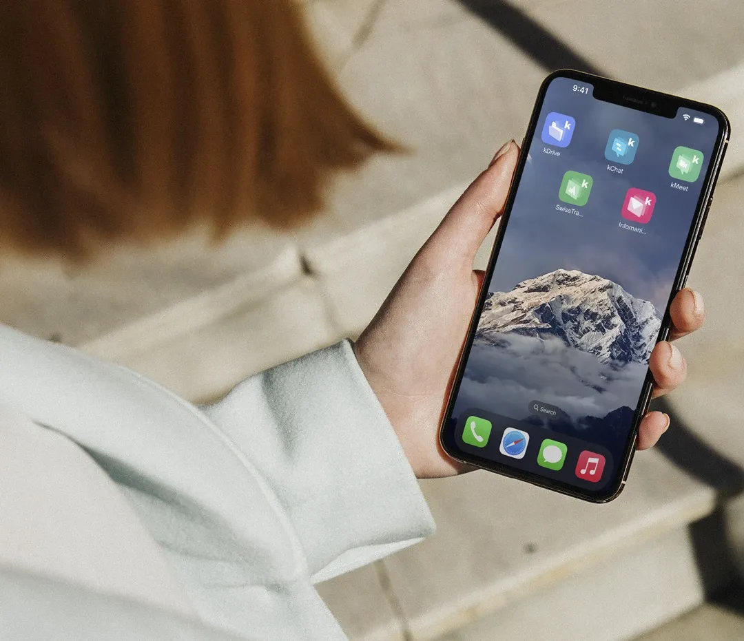 Photo d'une main tenant un smartphone affichant un écran d'accueil avec des icônes d'applications kSuite, sur un fond d'image de montagne enneigée.