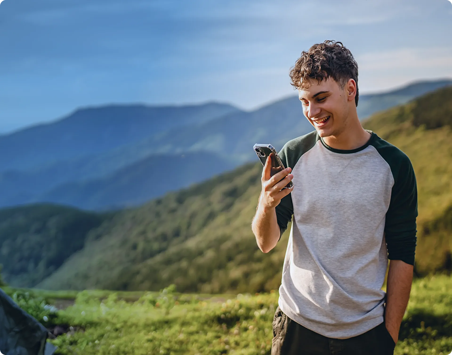 Un jeune homme portant un sac à dos, habillé d'un pull vert, utilisant son smartphone tout en se tenant dans un paysage naturel avec des collines et des arbres en arrière-plan.