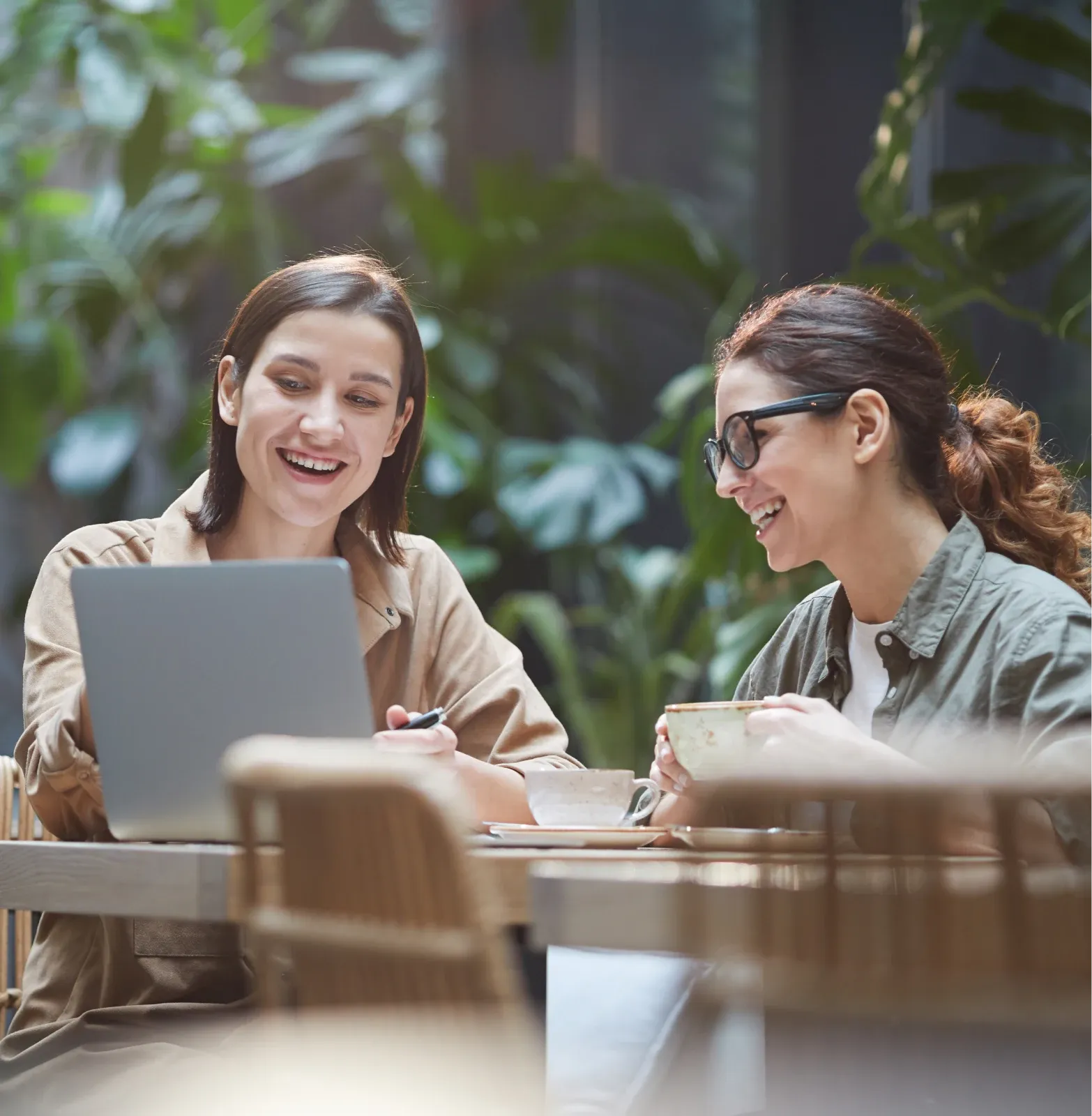 Deux femmes souriantes travaillant ensemble sur un ordinateur portable, assises à une table dans un espace lumineux et verdoyant, entouré de plantes.