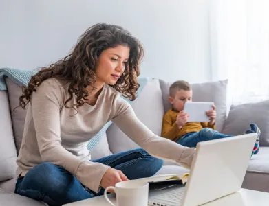Une femme travaillant sur un ordinateur portable dans un salon, avec un enfant jouant sur une tablette en arrière-plan, illustrant un environnement sécurisé et respectueux de la vie privée.