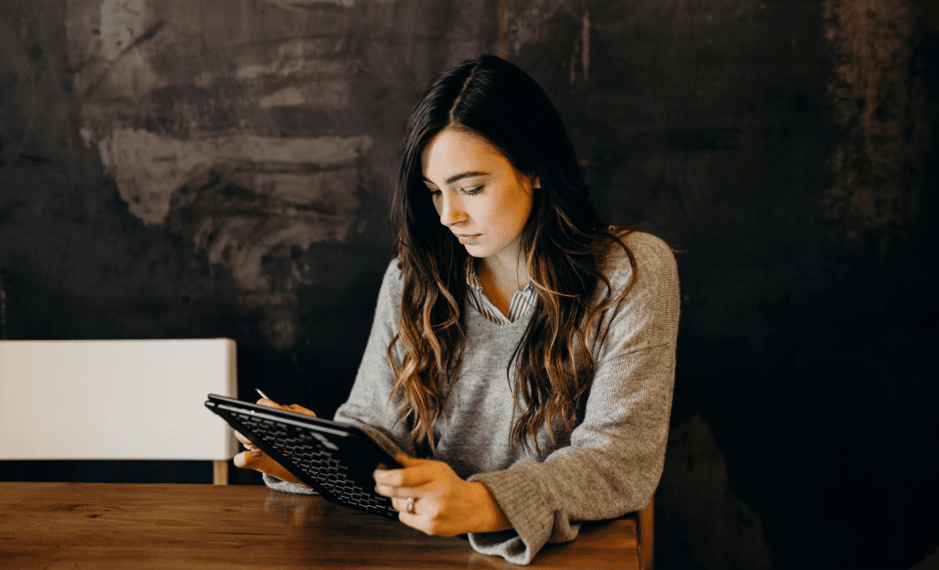 Une femme assise à une table en bois, concentrée sur l'utilisation d'une tablette dans un environnement sombre et chaleureux.